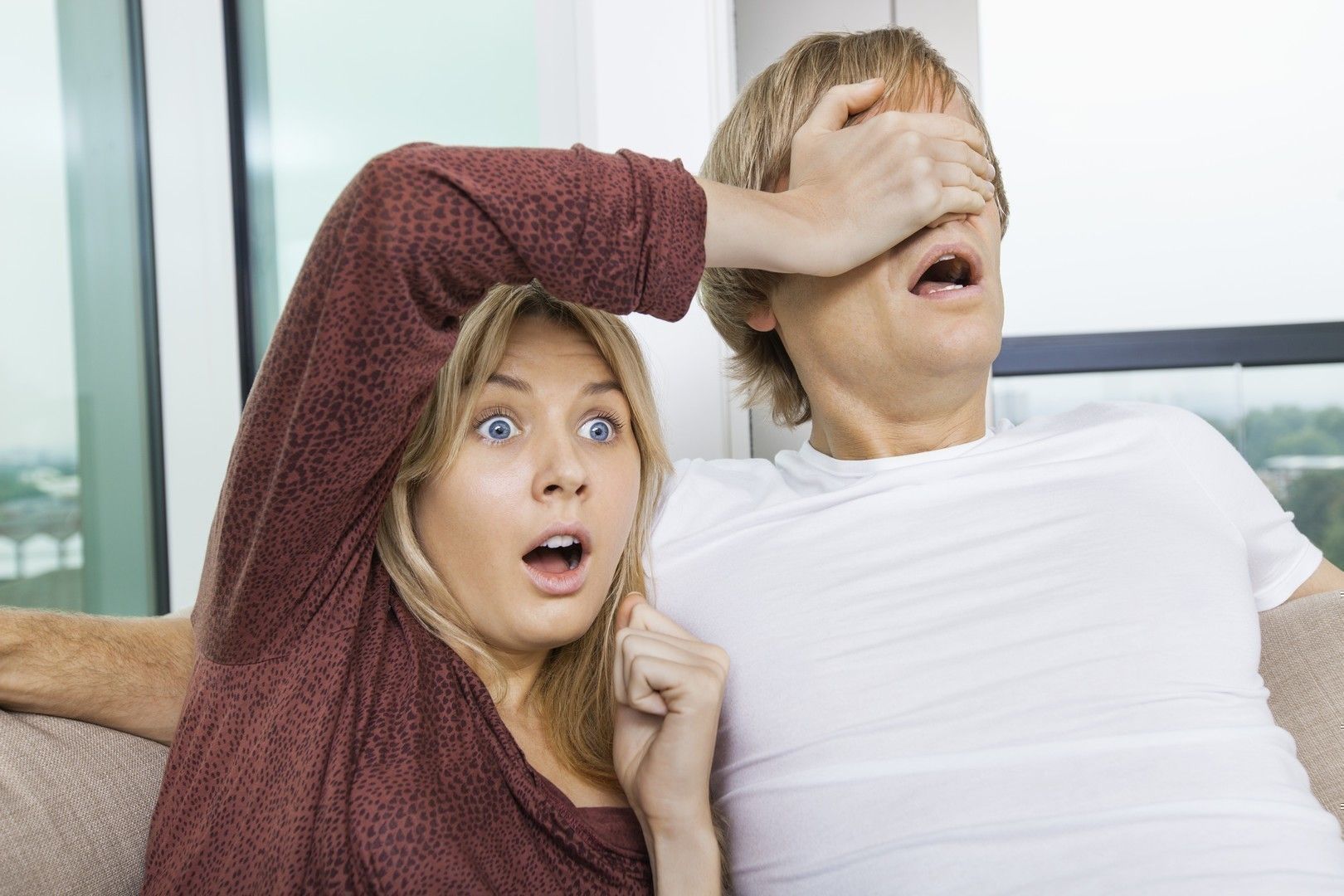 Shocked woman covering man’s eyes while watching TV at home