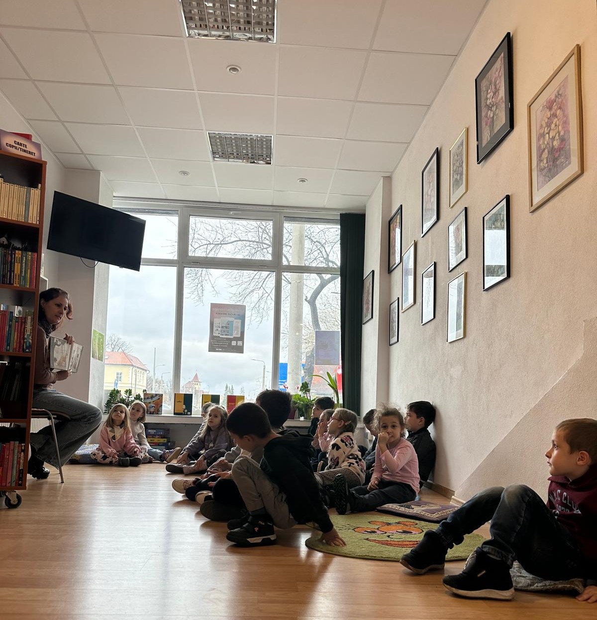 Preșcolari entuziasmați ascultând o poveste la bibliotecă ASTRA Sibiu, înconjurați de cărți colorate.