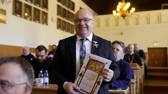Primarul Sadu onorat primește un plic de la un reprezentant al Arhiepiscopiei Sibiului în sala adunării