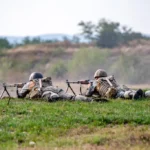 Soldați efectuând trageri cu muniție reală pe poligonul militar Poplaca, cu ținte în fundal.